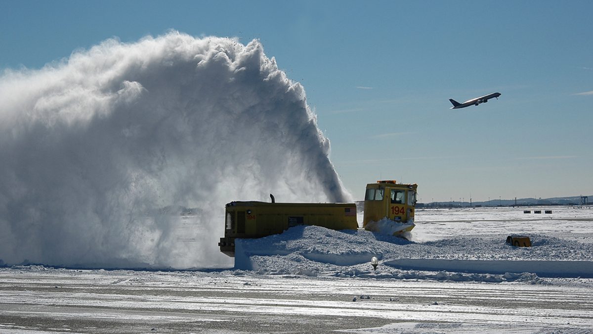 A snow blower throws a huge plume of snow across a runway at Boston Logan International Airport as a plane takes off in the background