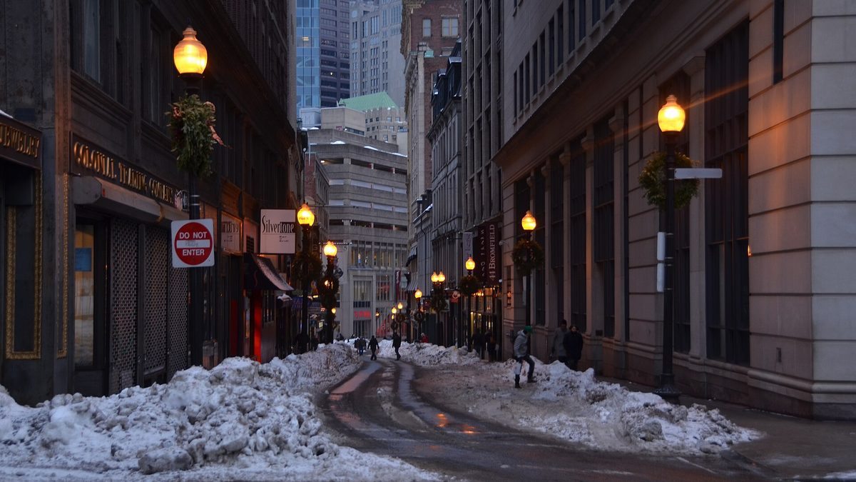 A snow-covered downtown Boston street at dusk with tall buildings and illuminated street lamps