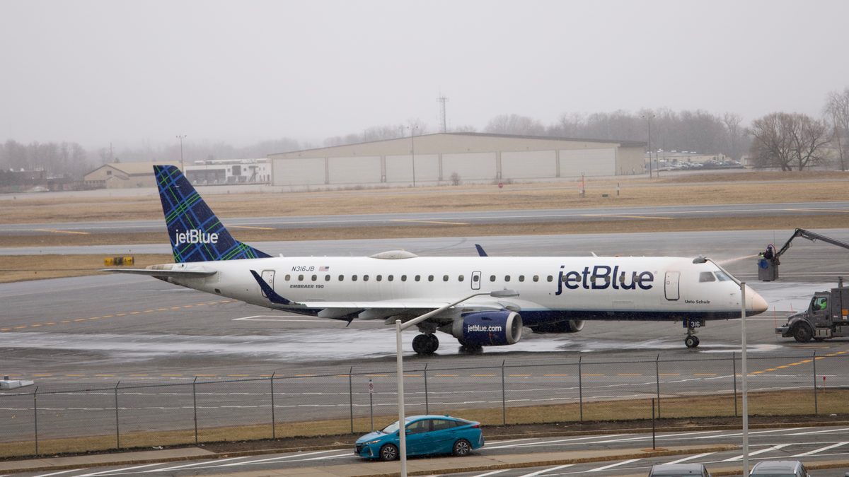 A JetBlue Embraer 190 being de-iced by a truck with a boom at Syracuse Hancock International Airport as snow begins to fall