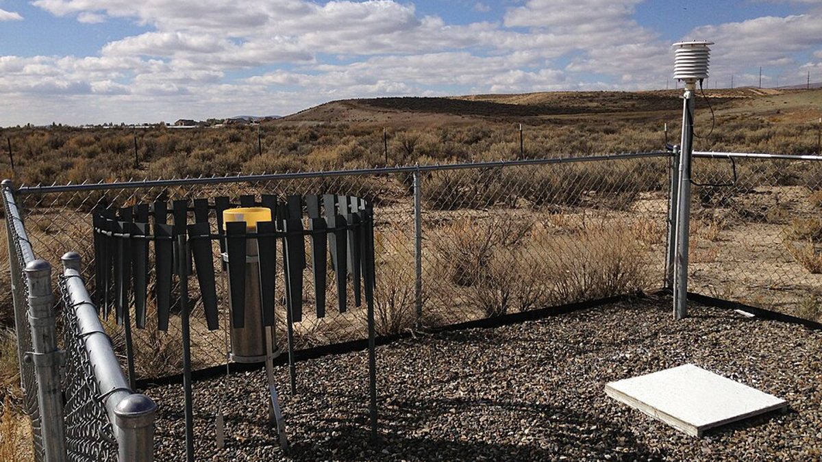 A fenced National Weather Service Cooperative Observer weather station with an alter-shielded precipitation gauge, a louvred thermometer shelter and a white snow board on gravel