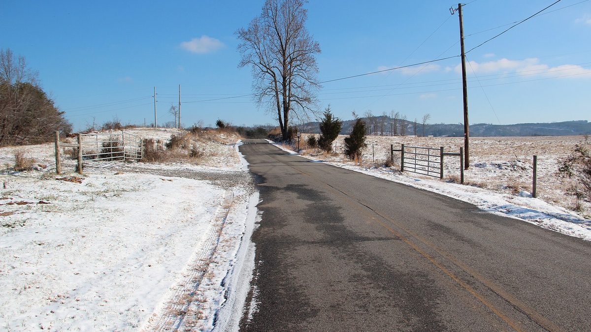 A rural road curving through north Georgia foothills with a thin dusting of snow on the grass verges and bare trees