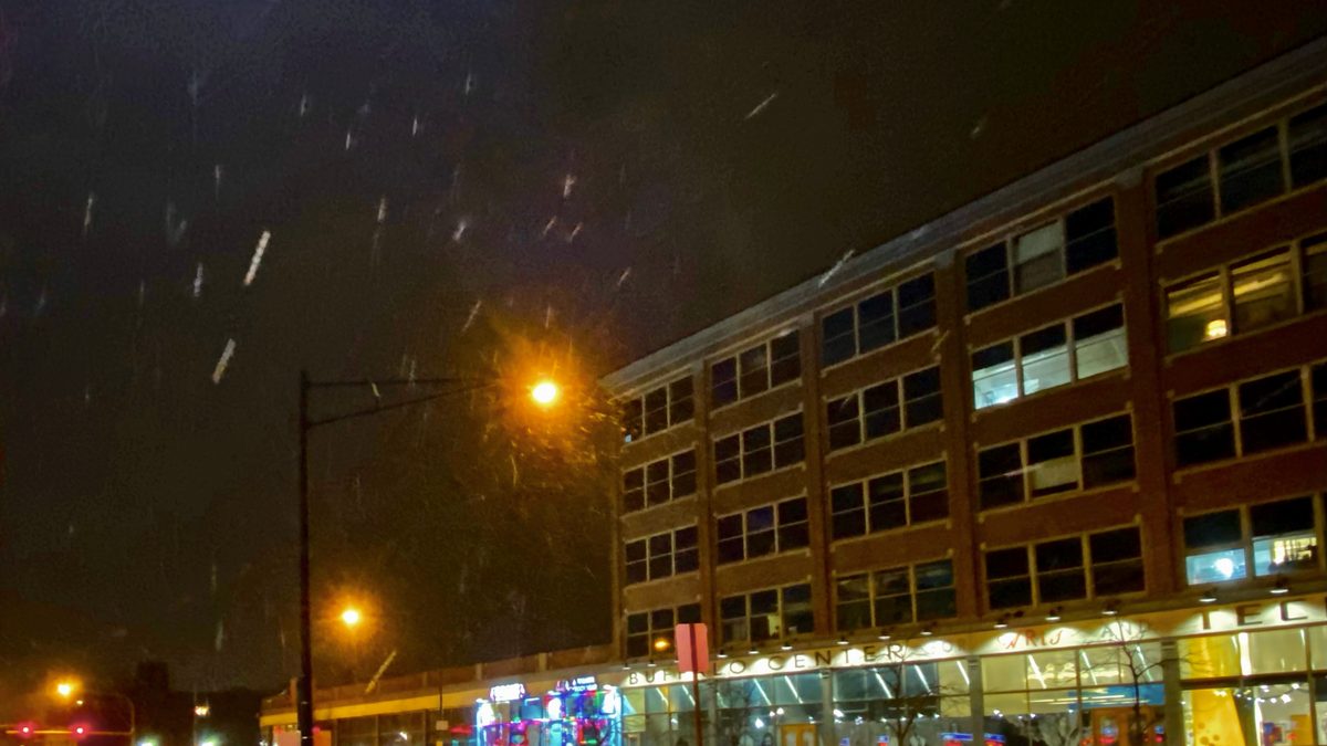 Snow falling at night over Main Street in Buffalo, New York, lit by a streetlight, with the Buffalo Center for the Arts building in the background